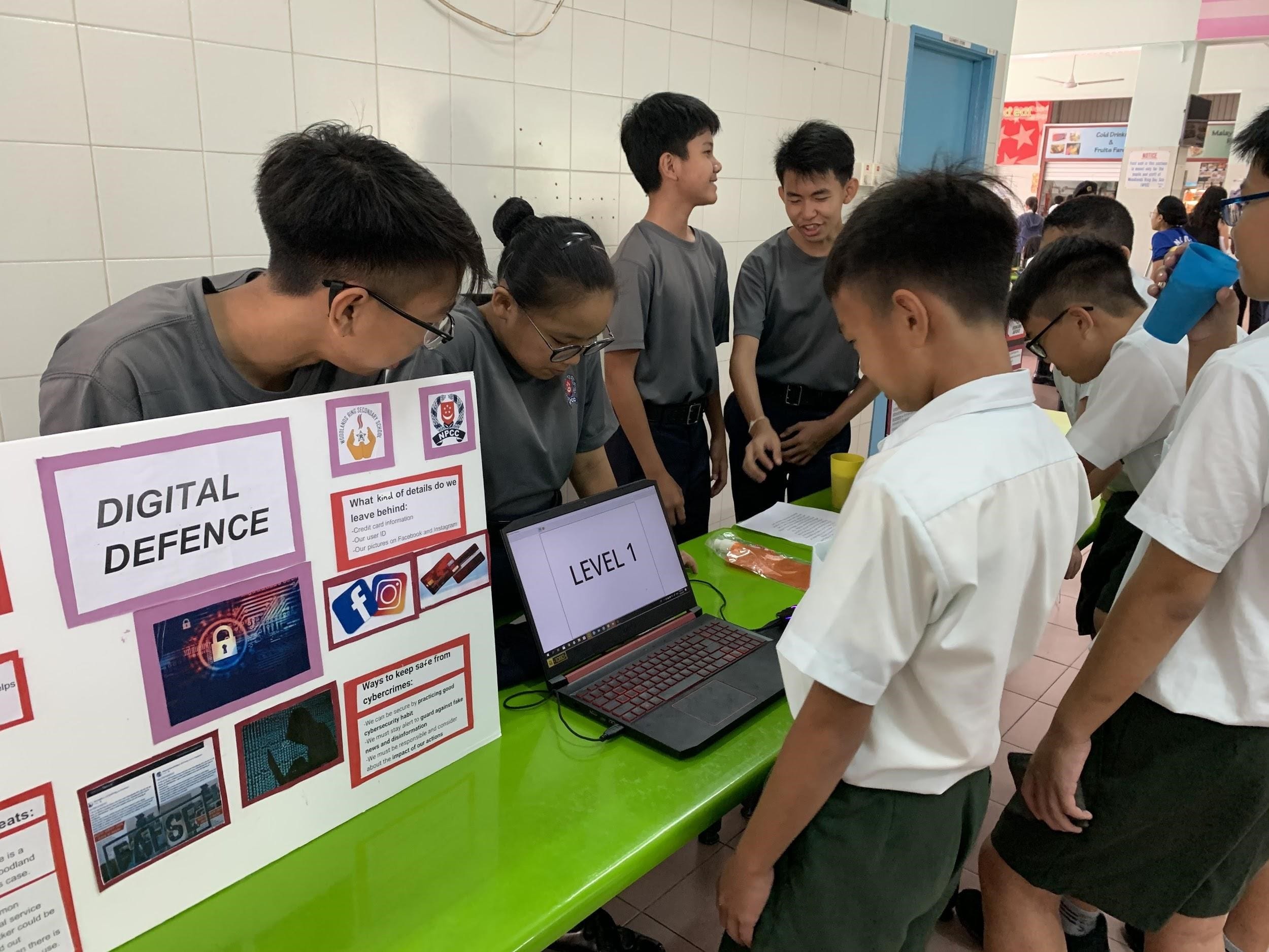 Students gather around a green table with a Digital Defence poster and a laptop showing Level 1 at a school event.